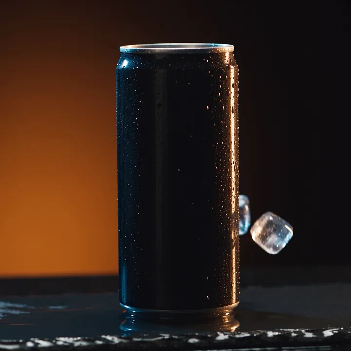 A black can beaded with condensation on a dark set, one warm rim light, an ice cube against it